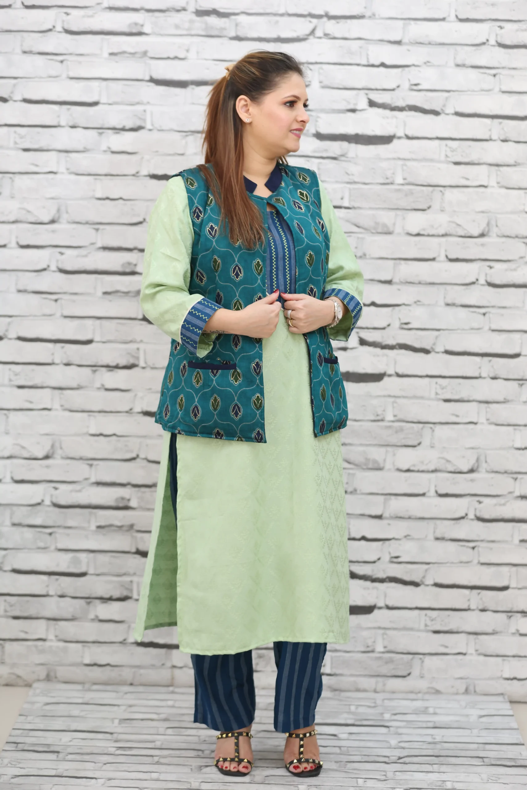 Woman wearing a sleeveless teal jacket with embroidered leaf motifs, crepe lining, and front pockets, styled over a light green kurta.