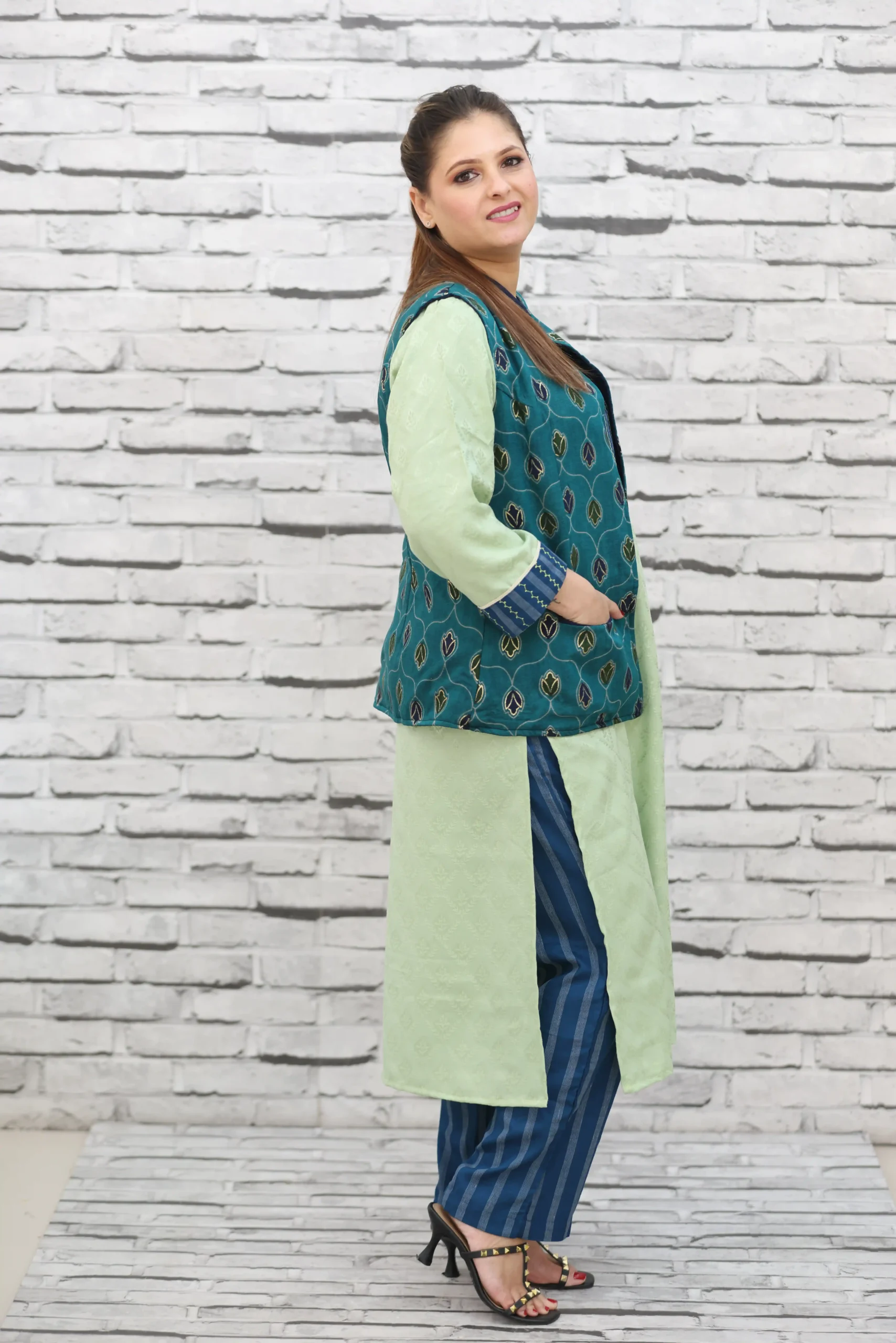 Woman wearing a sleeveless teal jacket with embroidered leaf motifs, crepe lining, and front pockets, styled over a light green kurta.