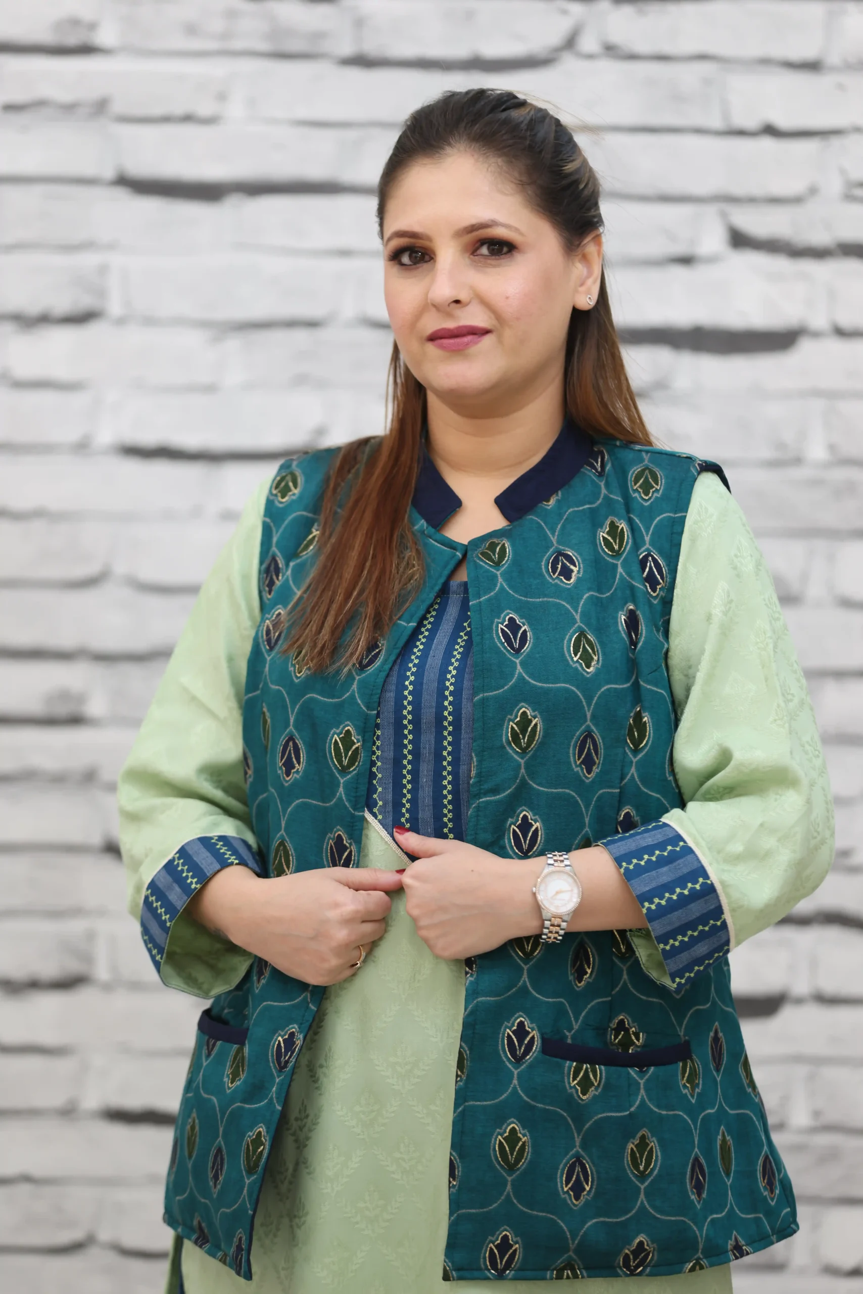 Woman wearing a sleeveless teal jacket with embroidered leaf motifs, crepe lining, and front pockets, styled over a light green kurta.